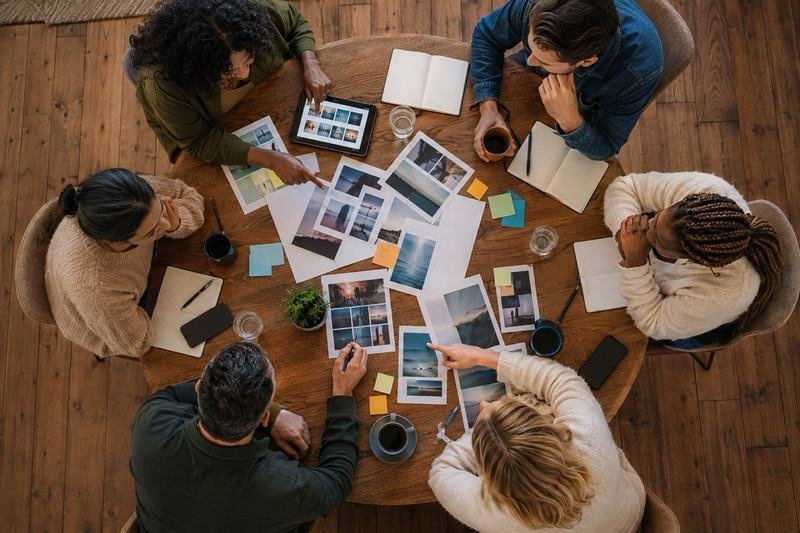 Seen from above, six people at a round wooden table sorting printed photographs and notes