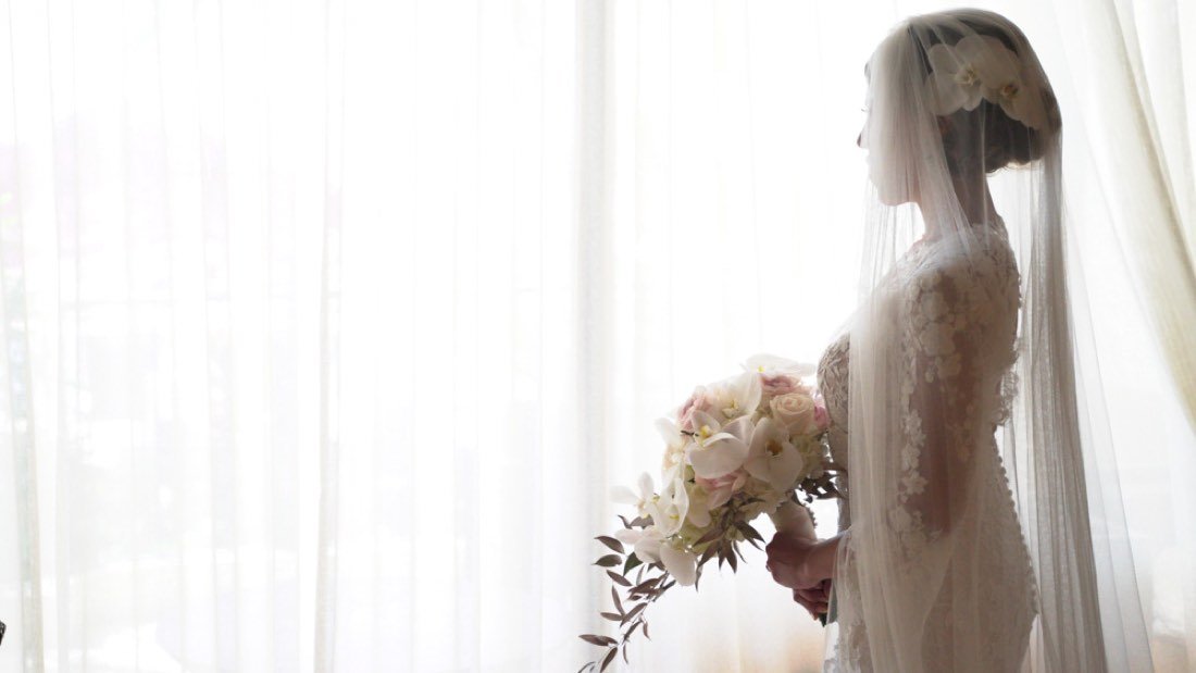 A bride backlit at a window, holding her bouquet